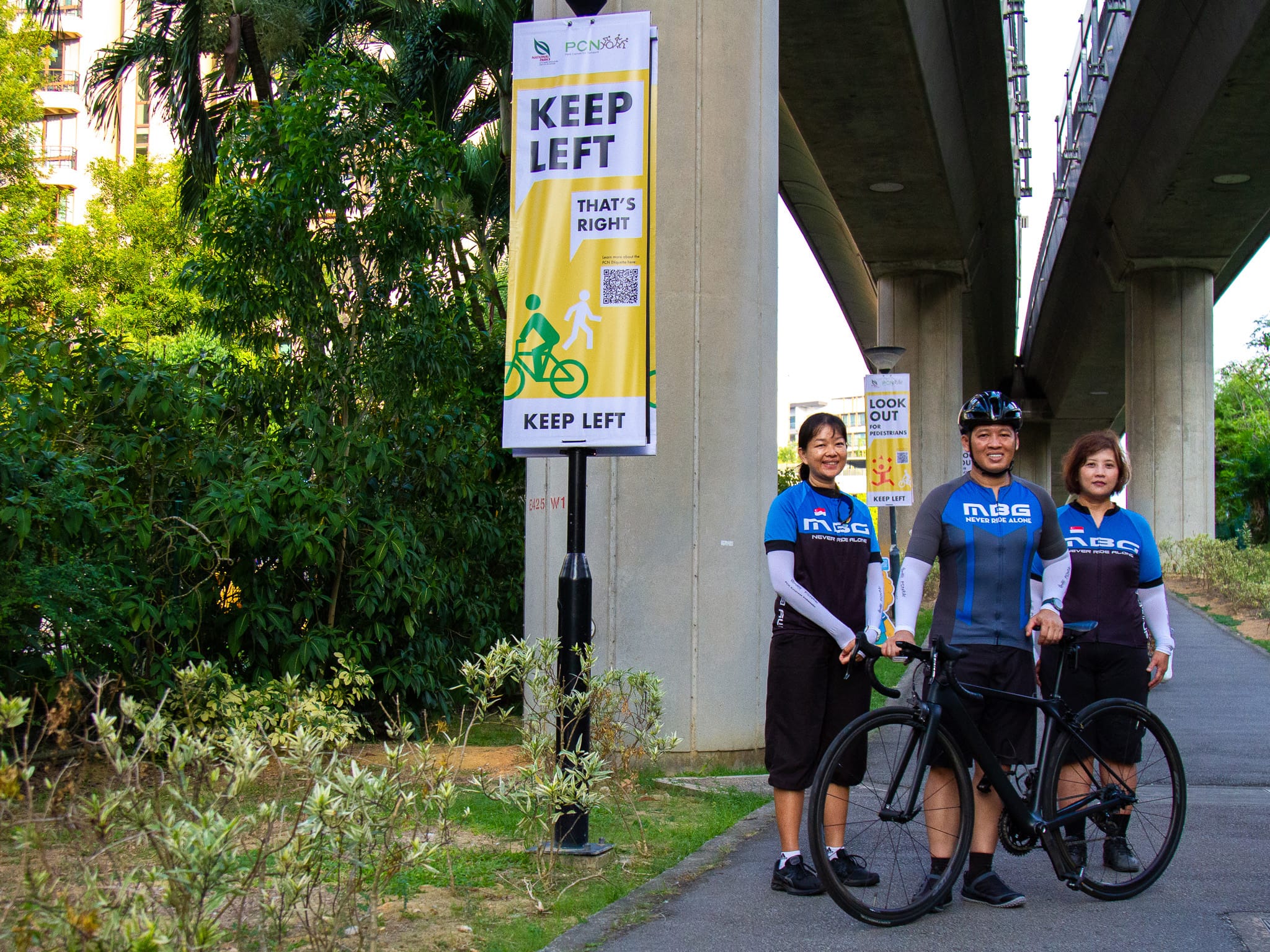Three cyclists in matching jerseys pose with a road bike by a Keep Left sign under an elevated walkway.